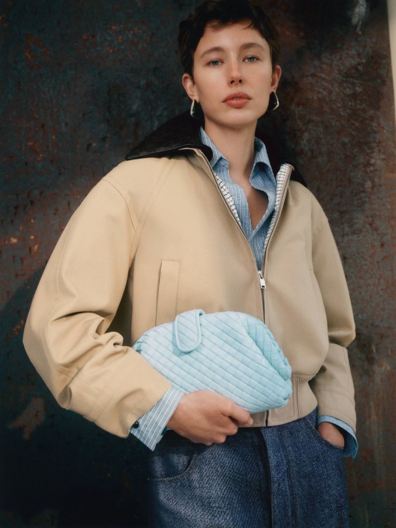 A young woman models a tan bomber jacket and jeans, holding a light blue quilted clutch, posing in front of a mottled brown backdrop.