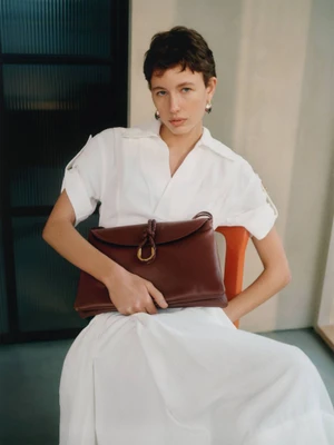 Model in a white dress holding a dark brown leather handbag against an interior architectural background