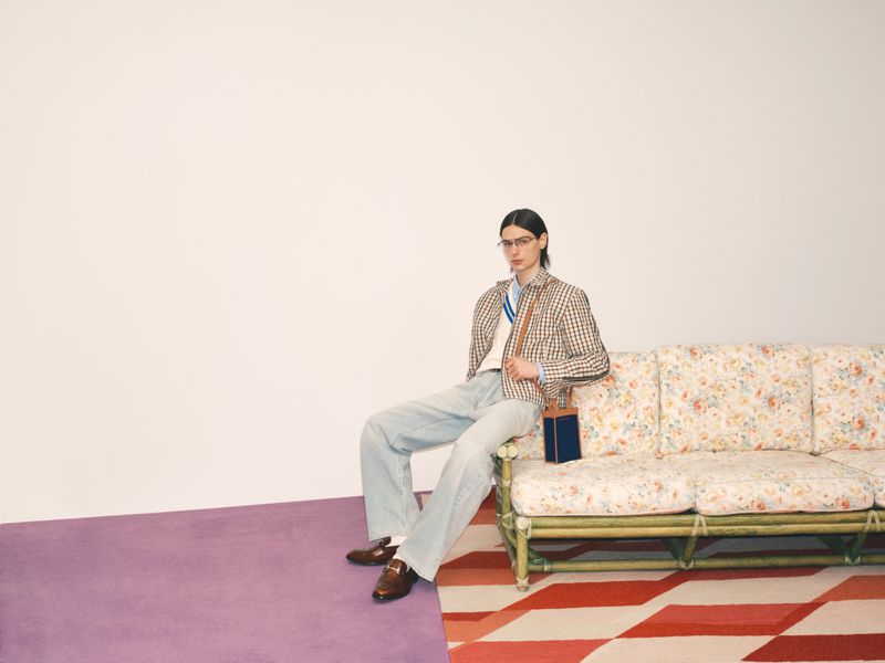 A slender adult in glasses sits on a floral couch wearing a blazer, light jeans, and loafers in a brightly lit studio fashion shot.