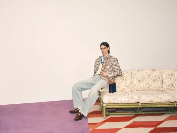 A slender adult in glasses sits on a floral couch wearing a blazer, light jeans, and loafers in a brightly lit studio fashion shot.