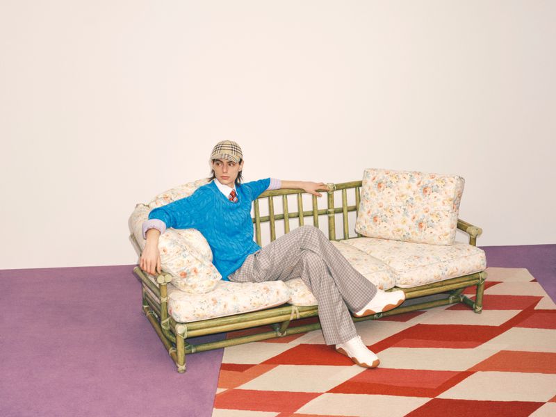 A young adult is sitting on a floral couch with a blue sweater, patterned pants, and a hat, on a carpeted floor against a white background.