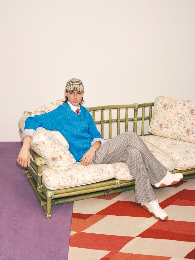Young adult poses on floral sofa in a checkered cap and pants, bright blue sweater, and white shoes against a white backdrop.