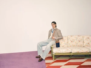 Male model in a plaid jacket and light wash jeans seated on a floral patterned sofa in a studio setting.
