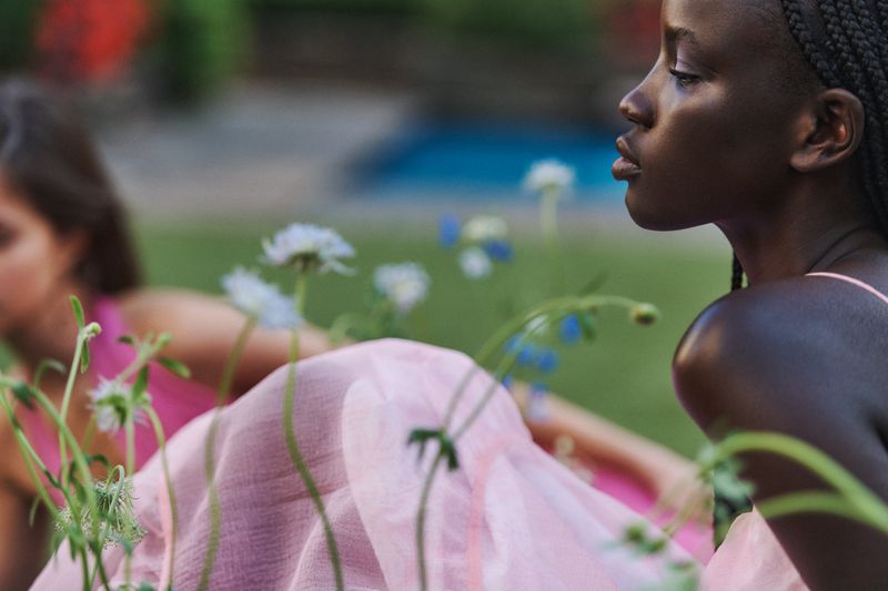 Two women sit in a garden with blooming flowers. One dark-skinned woman is in profile and wearing a pink dress. Second woman blurred.