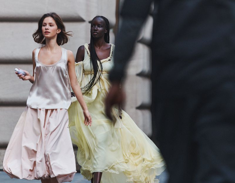 Two fashion models in flowing gowns walk on a city street. One wears a pale pink satin top and skirt, the other a light yellow dress.