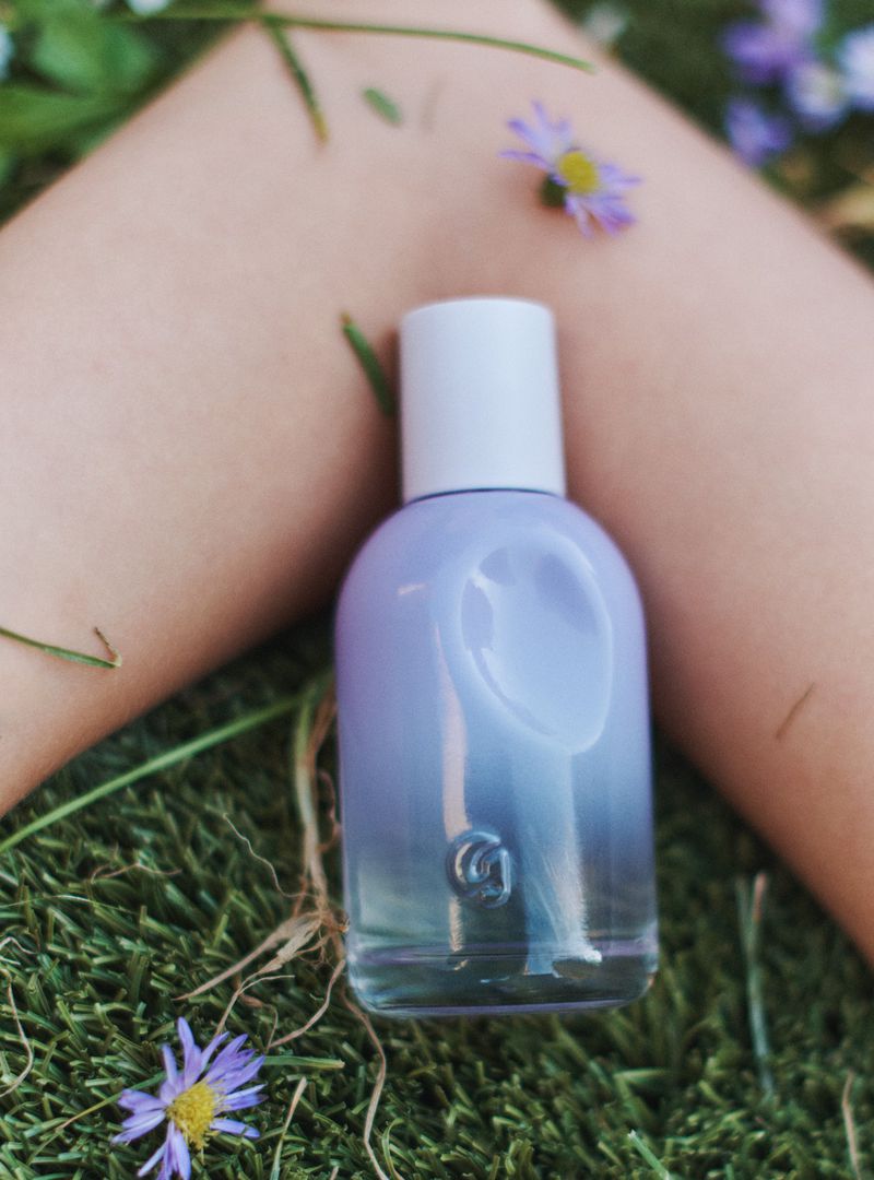 A bottle of perfume on a patch of green grass surrounded by small purple flowers. The bottle is light blue and lavender colored.