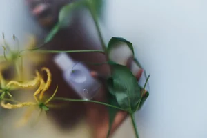 Close-up portrait of model Mayor Dutie holding a Glossier fragrance bottle amidst green foliage and yellow flowers.