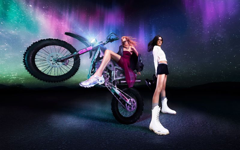 Two women pose with a motorcycle doing a wheelie in front of an aurora borealis backdrop in a fashion-forward editorial style photograph.