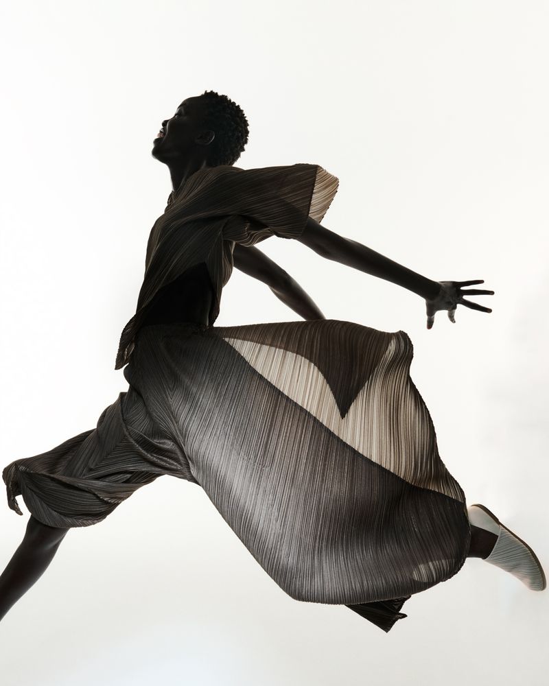 Silhouette of a slender, dark-skinned person in mid-air, wearing flowing fabric, gracefully leaping against a bright white background.