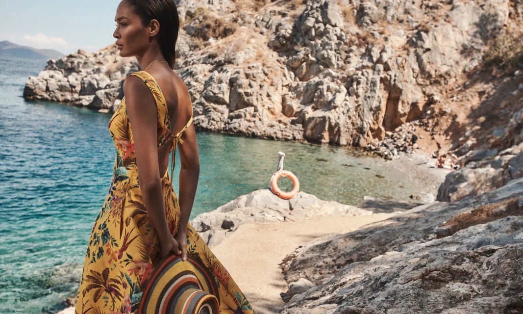 Woman in floral dress on rocky coast overlooking turquoise ocean. Wearing hat. Summer fashion, coastal scenery, and ocean backdrop.