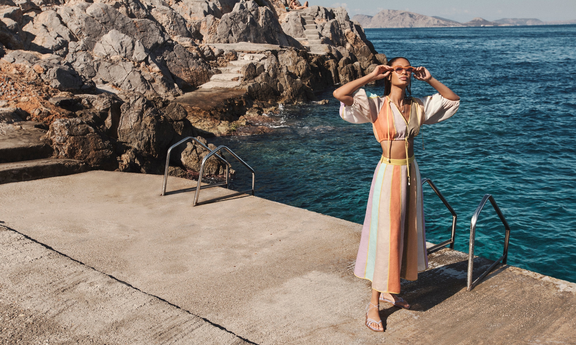 Adult woman in a colorful two-piece outfit and sunglasses poses on a concrete deck by the sea, with rocky cliffs in the background.
