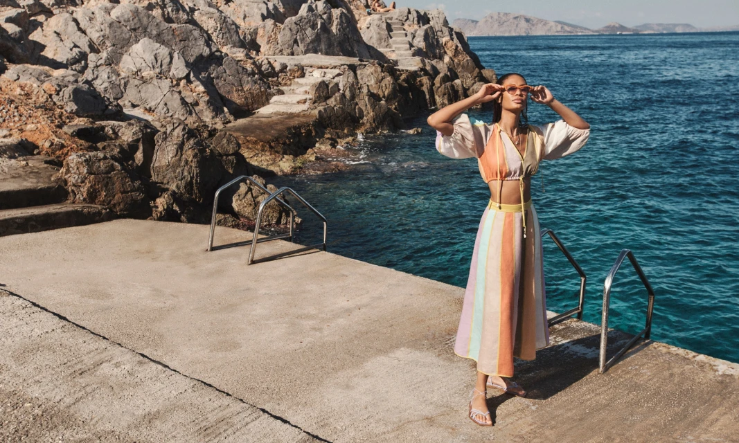 Adult woman in a colorful two-piece outfit and sunglasses poses on a concrete deck by the sea, with rocky cliffs in the background.