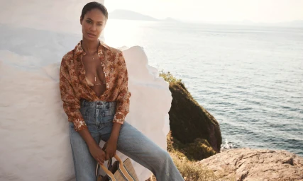 Model wearing a patterned shirt and blue jeans leaning against a white rock overlooking the ocean
