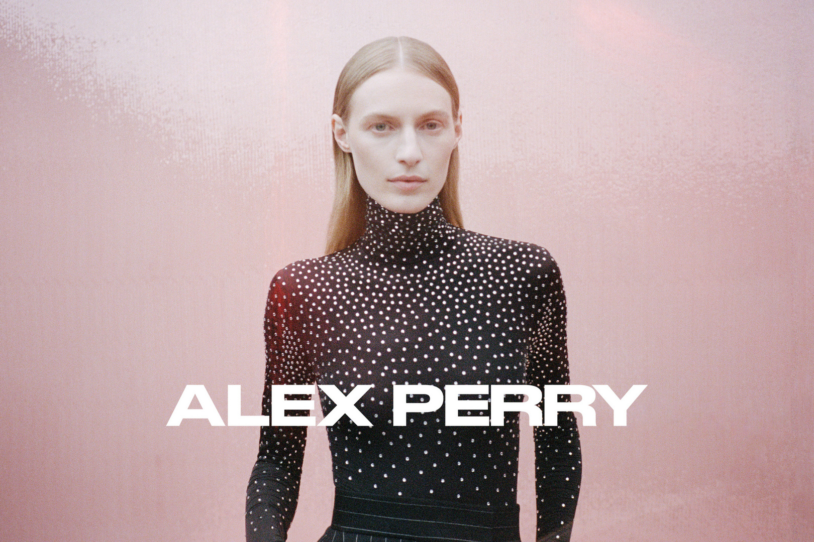 Fashion shot of a woman wearing a black long-sleeved turtleneck with silver embellishments posing in front of a pink background.