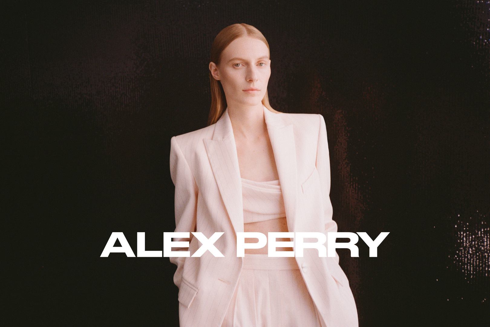 Fashion shot of woman in light pink suit jacket and pants against a dark background, featuring text overlay for Alex Perry brand.
