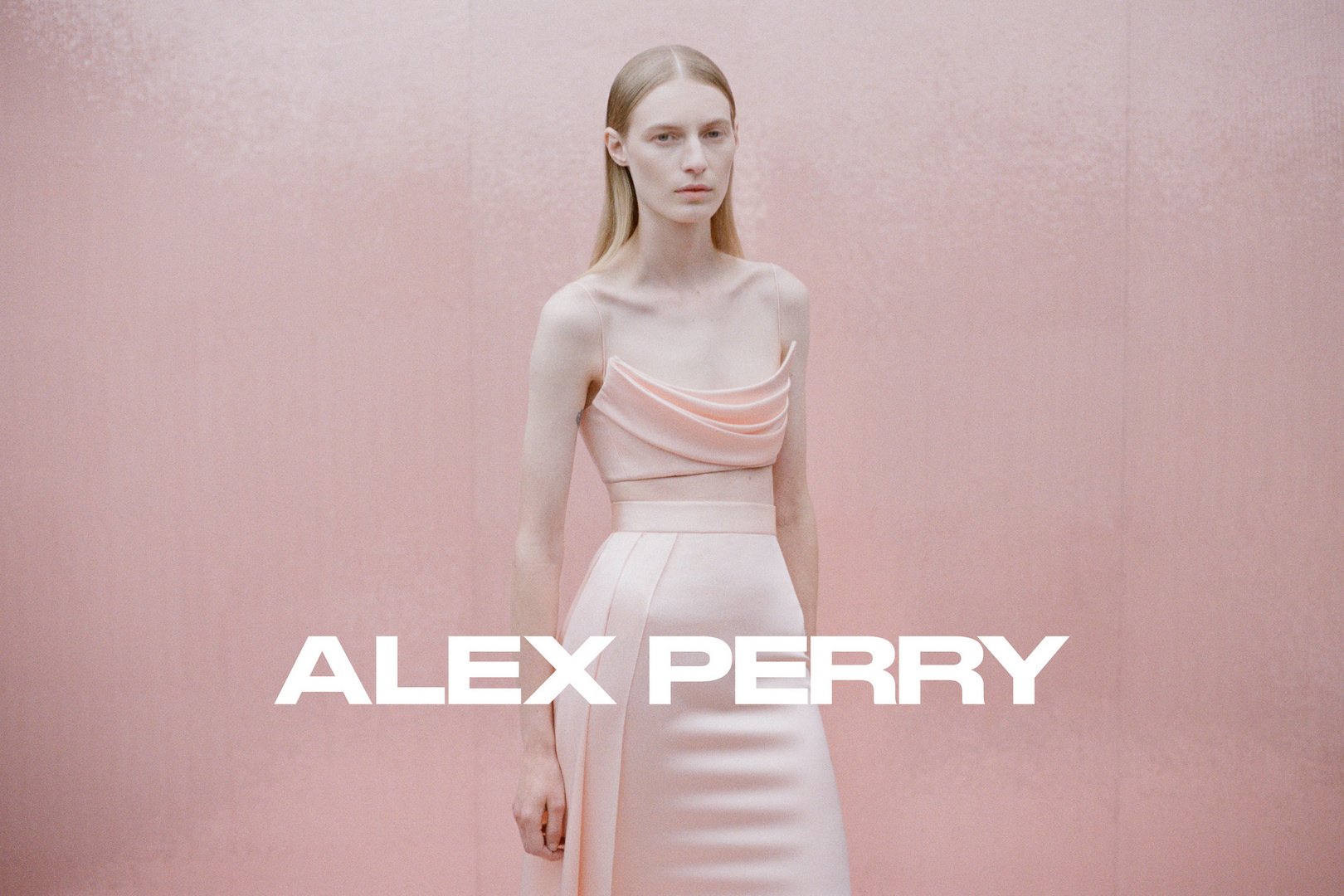 A slender female fashion model poses in a peach-colored two-piece dress against a pastel pink background, promoting Alex Perry designs.