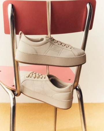 Two platform sneakers suspended by laces hang on the back of a retro metal chair, showcasing minimalist footwear design.