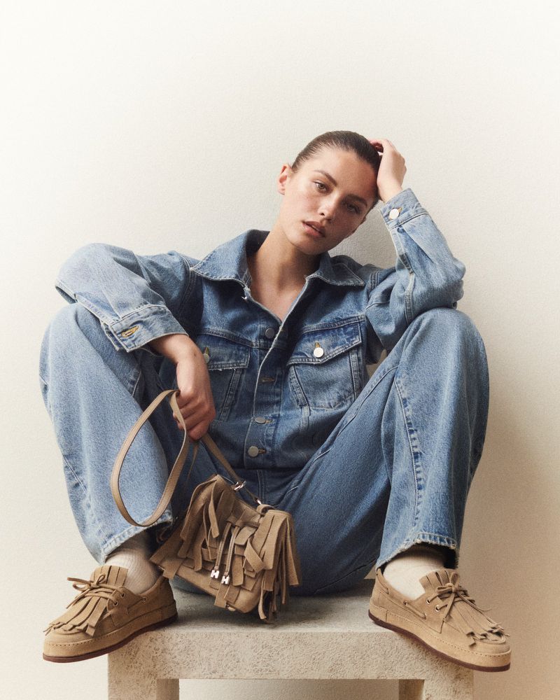 Model wearing a denim jacket and pants, holding a tan fringed handbag while seated on a stone block against a white backdrop.