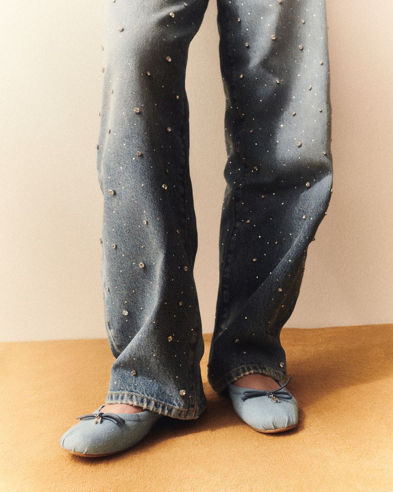 Close-up of lower body showing blue denim pants with jewel accents and light blue ballet shoes with ribbon on a tan floor and wall.