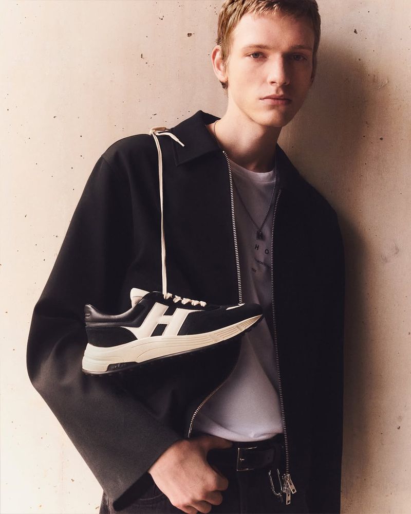 A young man wears a black jacket and holds a black and white leather sneaker with a white strap, looking at the camera.
