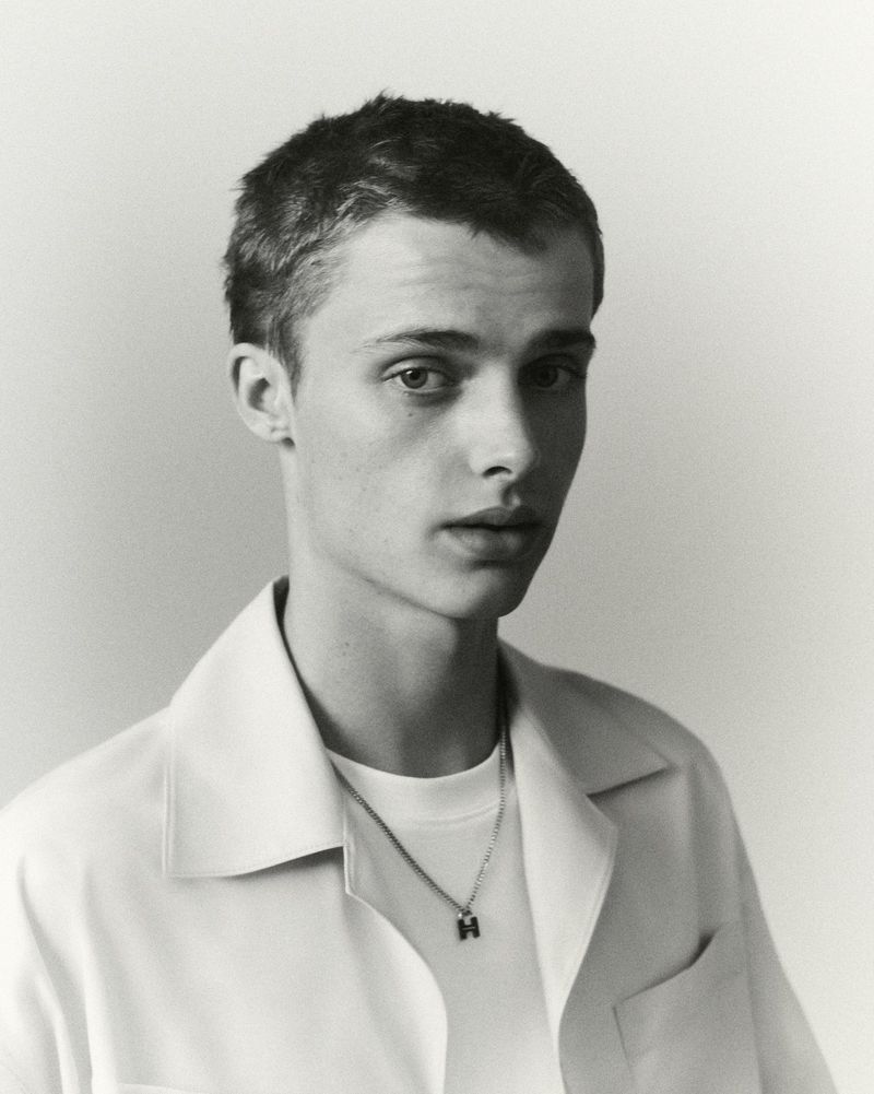 Portrait of a young fair-skinned man with short brown hair wearing a white collared shirt and 'H' pendant against a light background.