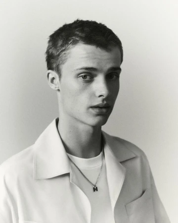Portrait of a young fair-skinned man with short brown hair wearing a white collared shirt and 'H' pendant against a light background.