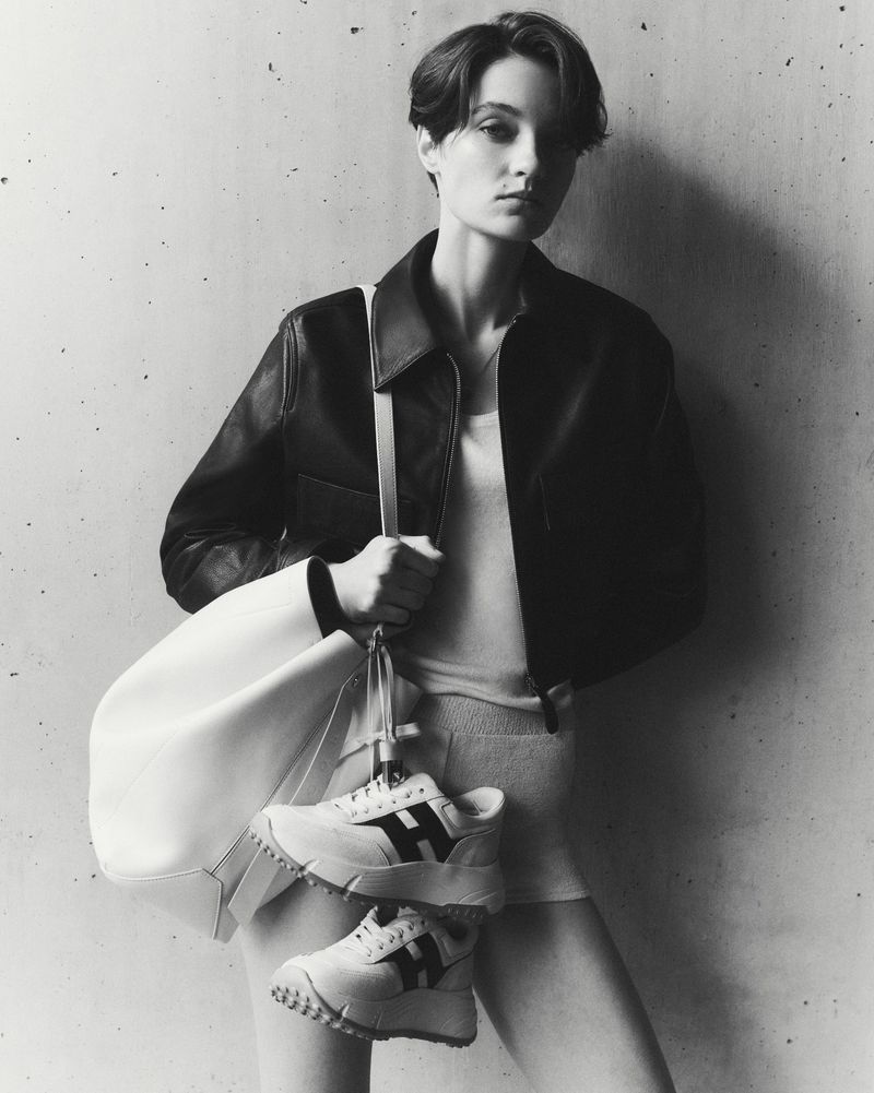 Model in dark leather jacket holding a white handbag with sneakers against a concrete wall