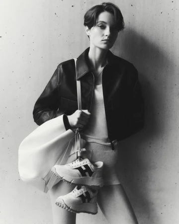 Model in dark leather jacket holding a white handbag with sneakers against a concrete wall