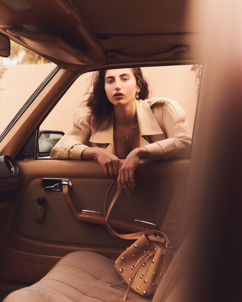 A woman in a light-colored coat leans out the window of a vintage car, with a tan studded purse on the tan car seat.