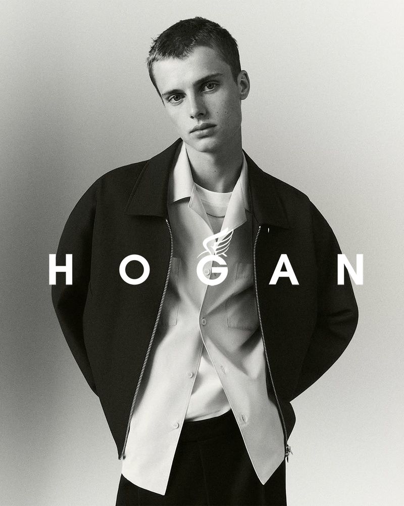 A young adult man in a dark jacket and light shirt poses in a studio against a gradient backdrop, showcasing fashion and style.