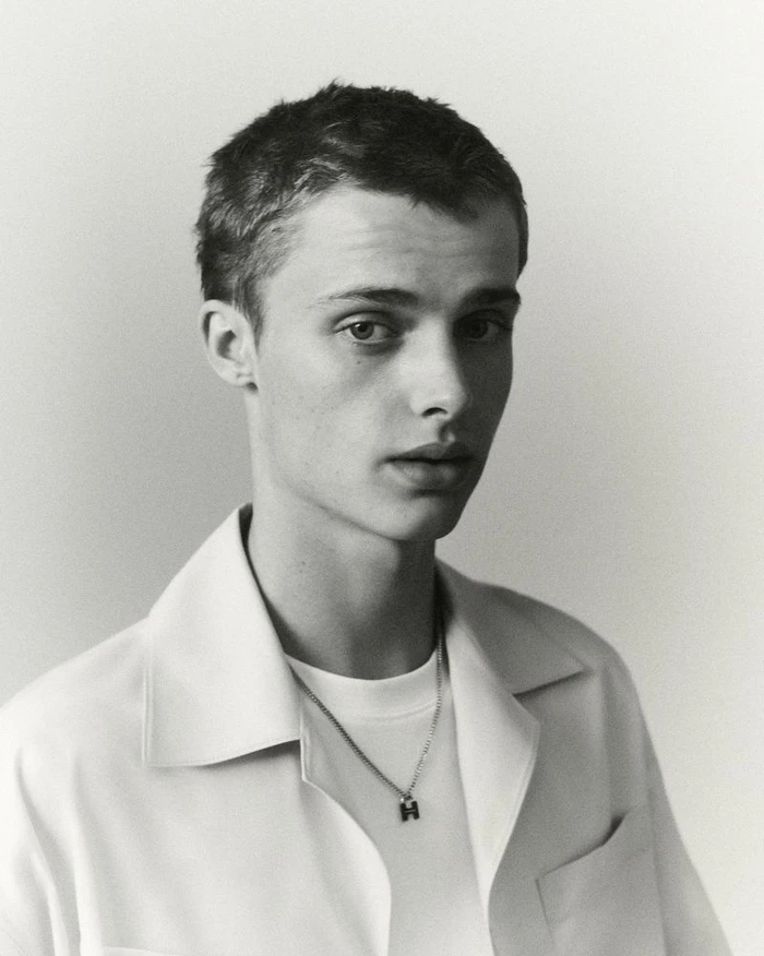 Portrait of a young male model wearing a white shirt and a pendant necklace against a plain light background