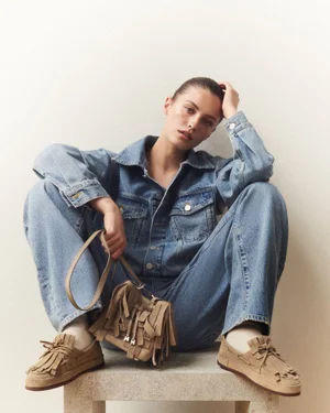 Model wearing a denim jacket and pants, holding a tan fringed handbag while seated on a stone block against a white backdrop.