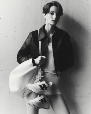 Model in dark leather jacket holding a white handbag with sneakers against a concrete wall