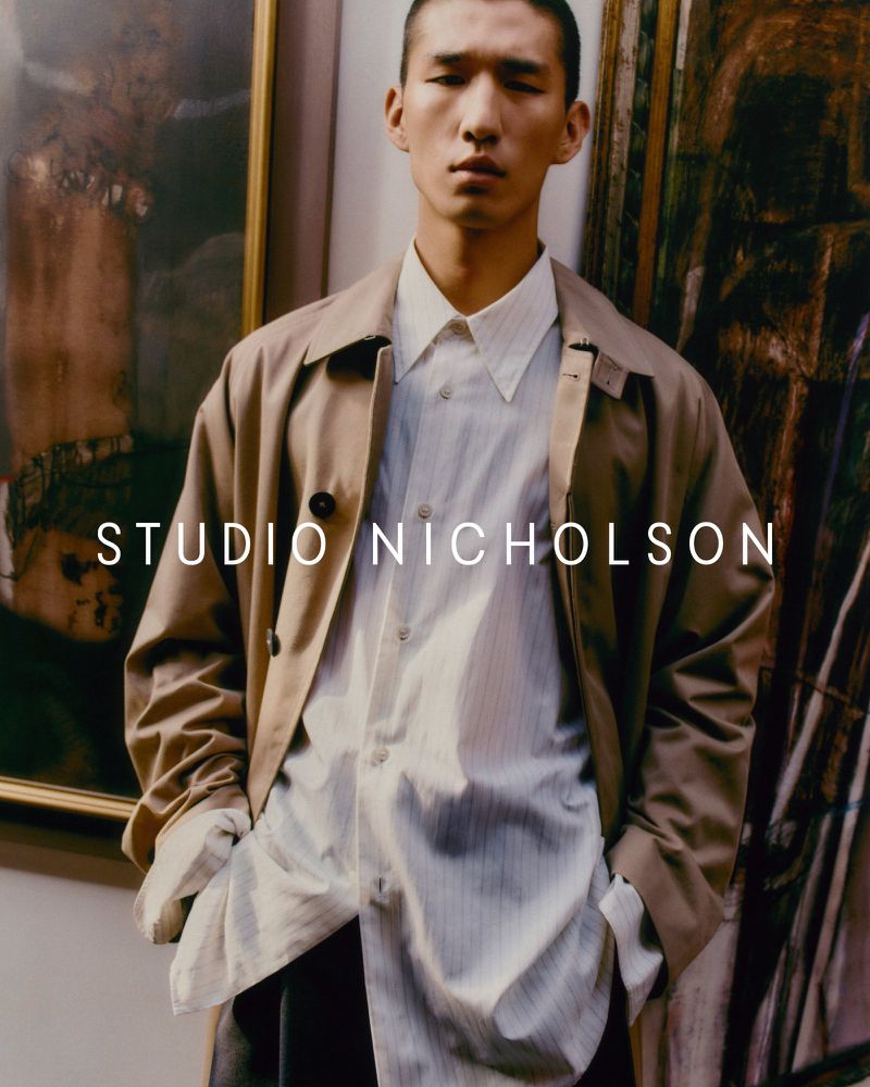 Portrait of an adult male wearing a button-down shirt and trench coat, posing in front of a framed painting in an indoor setting.