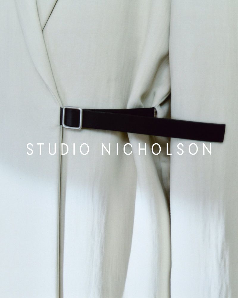 Studio Nicholson belted coat detail. Close-up on the black belt and buckle on a light beige coat in a neutral indoor setting.