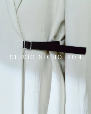 Studio Nicholson belted coat detail. Close-up on the black belt and buckle on a light beige coat in a neutral indoor setting.