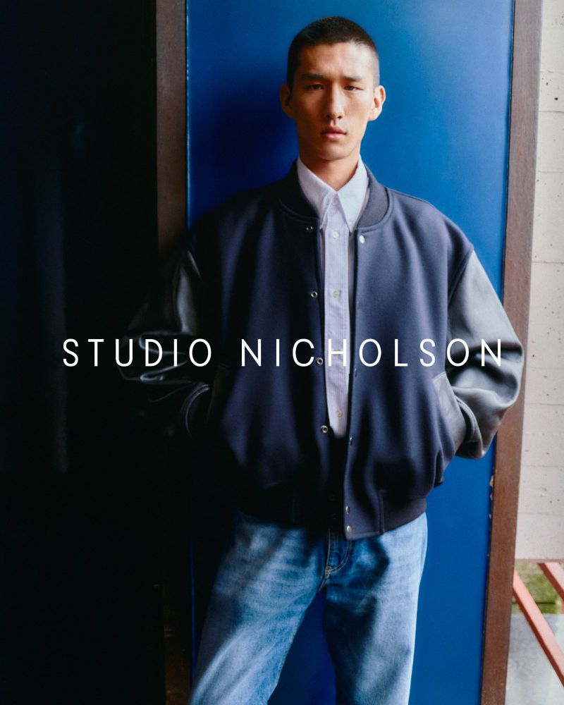 A stylish young adult man poses indoors wearing a navy bomber jacket, white collared shirt, and blue jeans against a blue wall.
