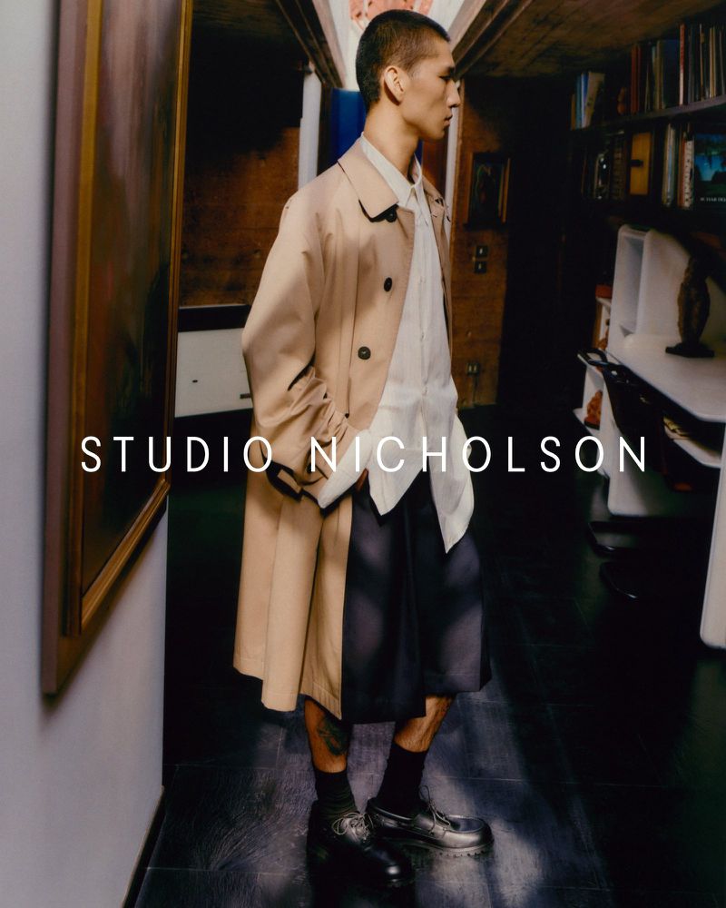 Studio Nicholson ad featuring a stylish Asian male model in a tan trench coat, white shirt, and dark shorts against an indoor setting.