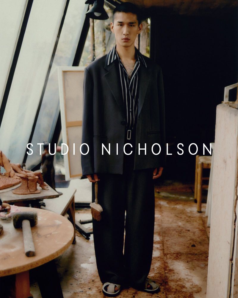 Adult male fashion model wearing dark suit, sandals, and striped shirt stands in a light-filled studio space with art supplies.