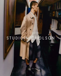 Man wearing a camel trench coat over a white shirt standing in a dimly lit hallway with artwork and bookshelves.