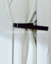 Close-up of a light beige garment secured with a dark black belt featuring a silver square buckle, with Studio Nicholson text overlay.