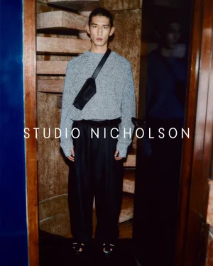 Model standing in front of a spiral staircase wearing a grey sweater and black crossbody bag for Studio Nicholson.