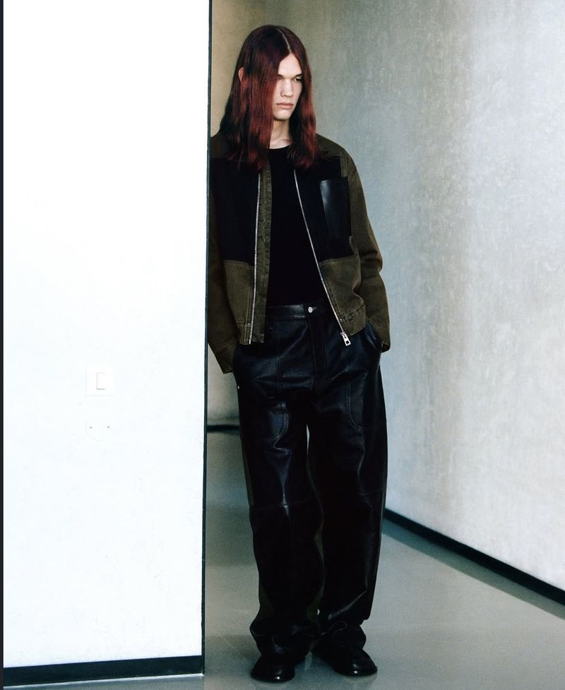 A person with long, reddish-brown hair poses in front of a corner wearing a black shirt, olive jacket, and baggy black leather pants.