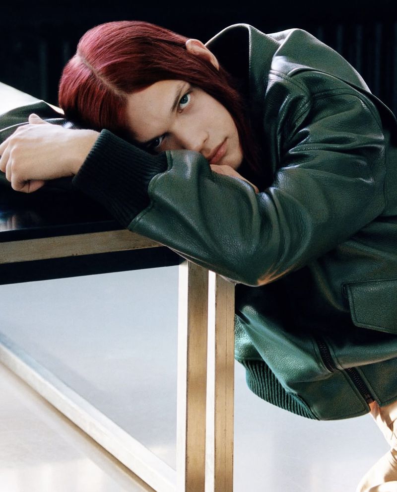 Portrait of an adult male with long auburn hair resting on a black table, wearing a green leather jacket with a neutral expression.