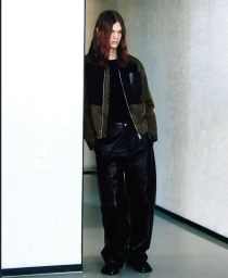 Model with long reddish hair wearing an olive jacket and black pants standing by a white wall