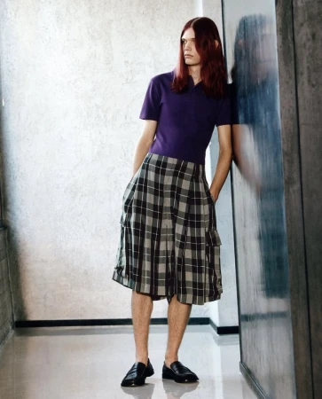 Model with long reddish-brown hair wearing a purple polo shirt and plaid pleated shorts leaning against a textured wall.