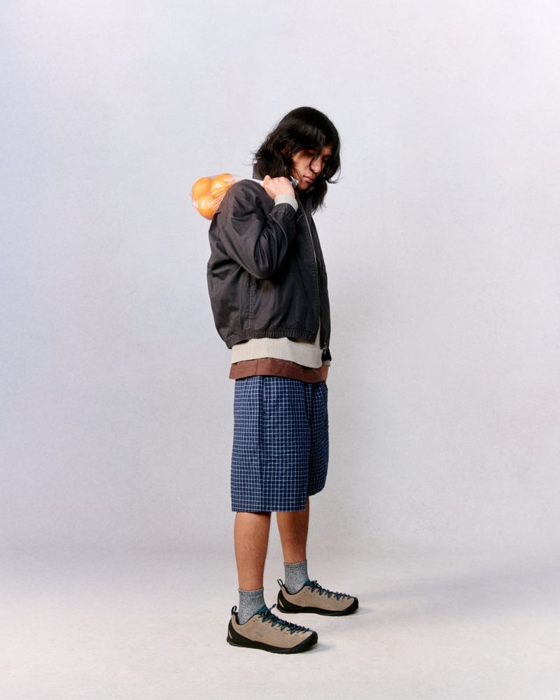 A long-haired person in a bomber jacket and shorts carries produce on their shoulder against a solid white backdrop in a studio setting.