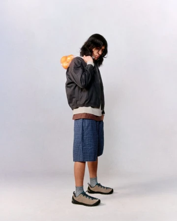 A long-haired person in a bomber jacket and shorts carries produce on their shoulder against a solid white backdrop in a studio setting.