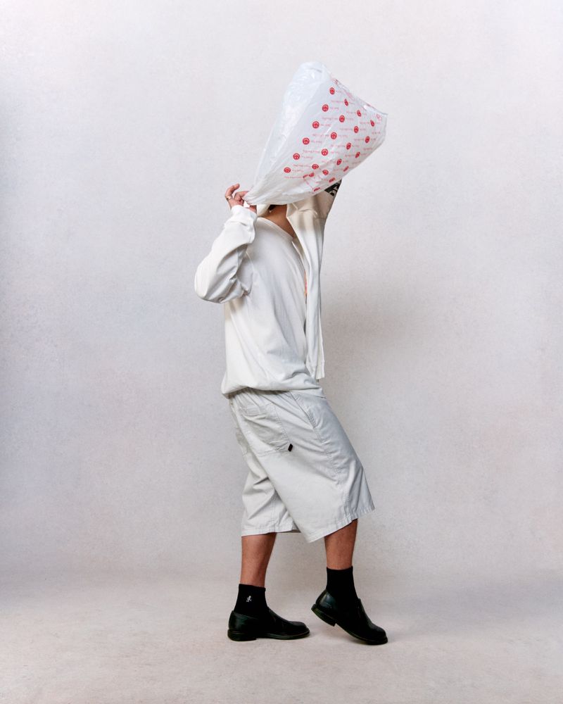 Person wearing a white outfit and black shoes with a plastic bag over their head stands in front of a plain white background.