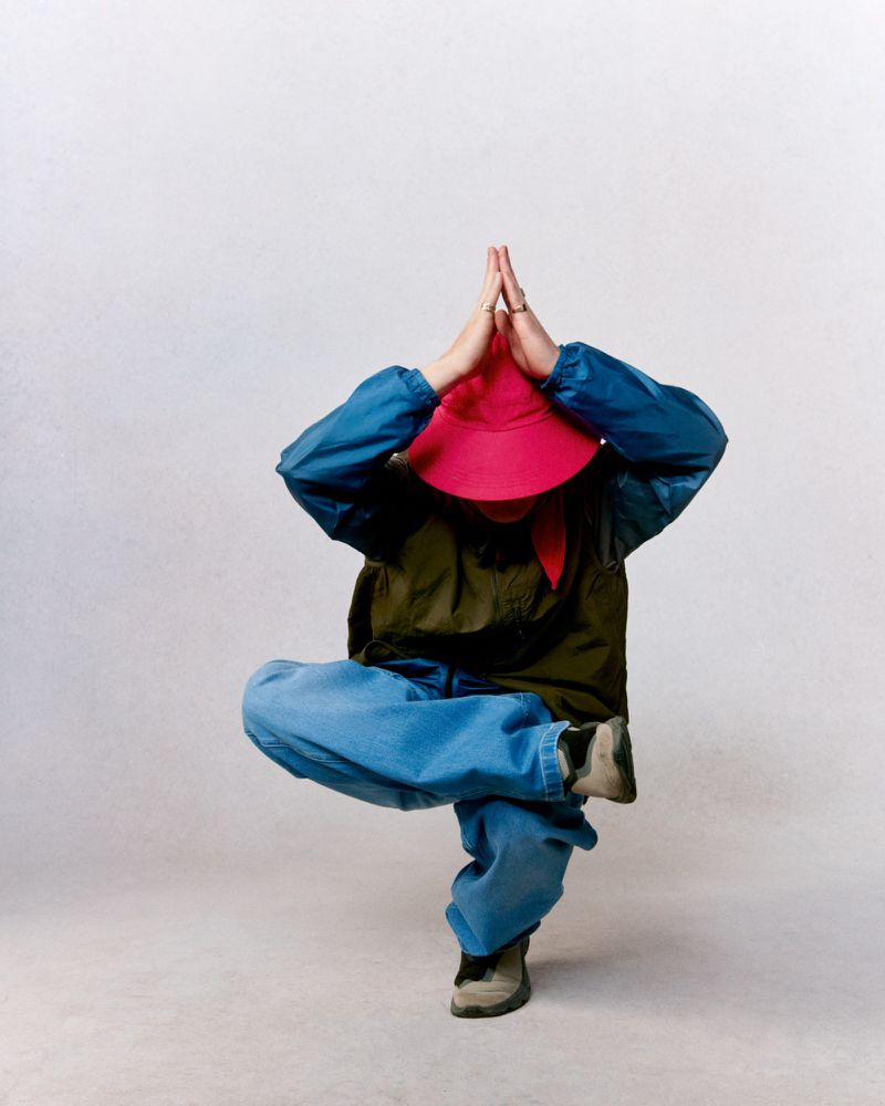 Person in balancing yoga pose wearing blue jacket, pants, green vest and red hat against white background in studio shot, overhead angle.